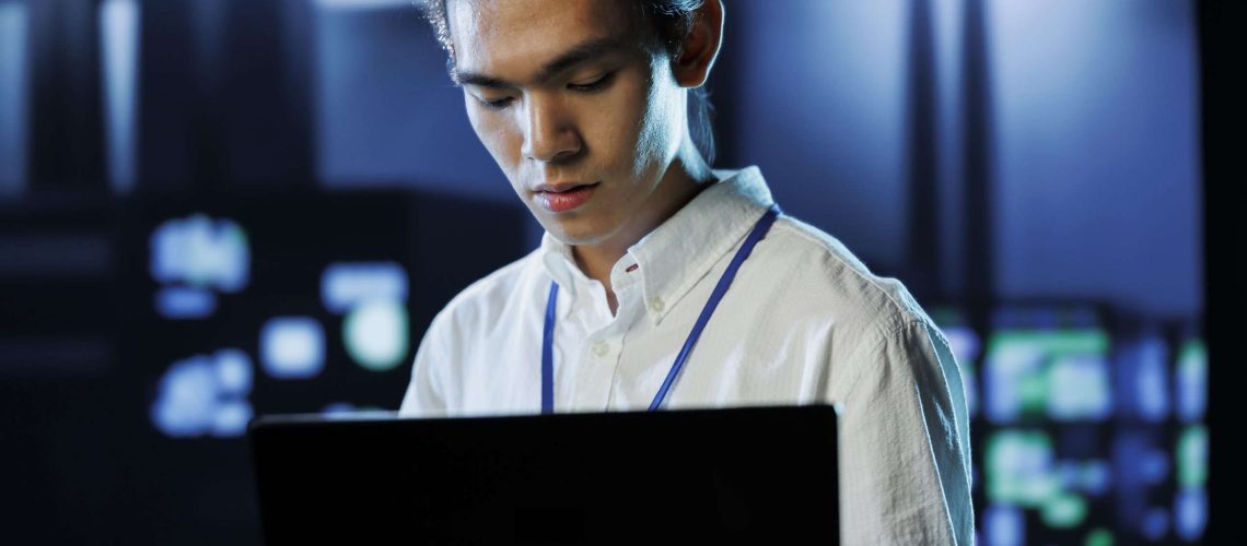 Dara center manager running code and programming on laptop, troubleshooting and upgrading high tech facility designed to accommodate rows of fully operational server supercomputers